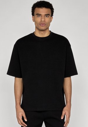 Mężczyzna z krótkimi, kręconymi włosami, ubrany w czarną teksturowaną koszulę z krótkim rękawem i dopasowane czarne spodnie, stojący na jednolitym tle.