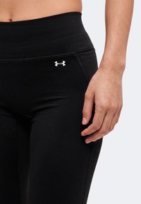 Gros plan d’une personne portant des leggings noirs Under Armour avec une taille haute et le logo sur la hanche, montrant sa main reposant à côté.