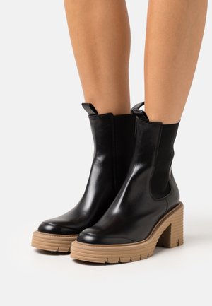 Bottes à cheville en cuir noir avec semelles épaisses et talons beiges portées sur des jambes nues sur un fond blanc.
