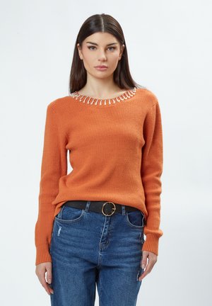 Pull en maille côtelée orange avec un col orné de bijoux, manches longues et coupe décontractée, associé à un jean en denim bleu et une ceinture noire.