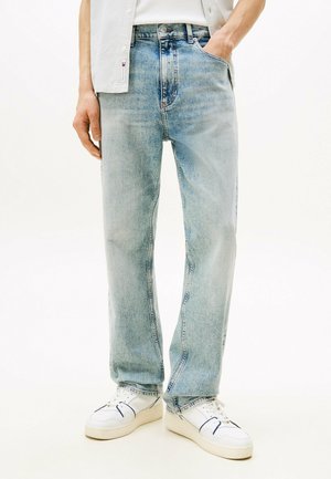 Mann trägt hellblaue Jeans mit geradem Bein, eine Hand in der Tasche und weiße Sneaker mit blauen Akzenten vor weißem Hintergrund.