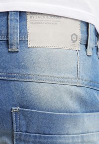 Sininen farkkuhousu, jossa on kulunut pinta. Housuissa on vahvistettu vyötärö, takatasku sekä vaaleasta kankaasta valmistettu etiketti.