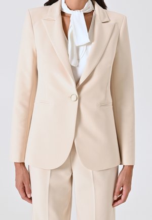 Kvinde iført beige skræddersyet blazer med enkelt knap og matchende bukser, kombineret med hvidt tørklæde bundet omkring halsen.