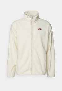 Krämfärgad fleecejacka med hög krage, hel dragkedja och en liten röd Nike-logotyp broderad på vänster bröst. Slät textur.