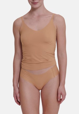 Nainen pukeutuneena beigeen saumattomaan kamisolaan ja yhteen sopiviin korkeavyötäröisiin alushousuihin, seisoo yksiväristä vaaleaa taustaa vasten.