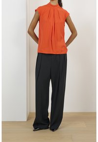 Blouse rouge à manches courtes avec des plis, associée à un pantalon noir taille haute à plis et des talons noirs à bout pointu, sur fond neutre.