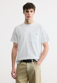 Νεαρός άνδρας που φοράει άσπρο κοντομάνικο μπλουζάκι με τσέπη στο στήθος και μπεζ τζιν, στέκεται μπροστά από λευκό, απλό φόντο.