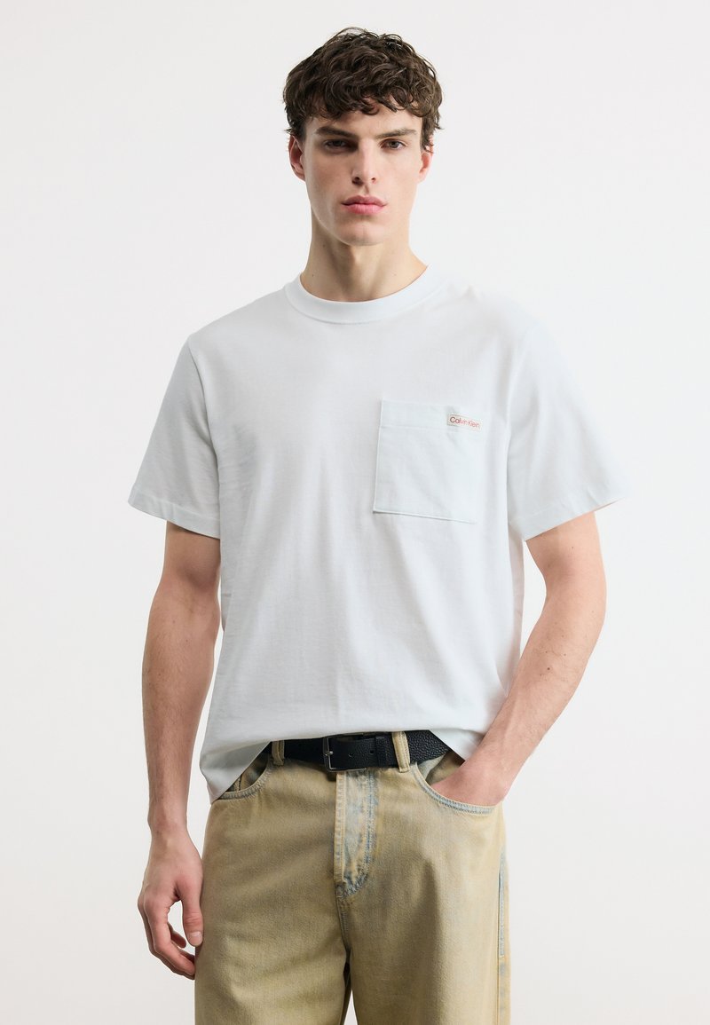 Νεαρός άνδρας που φοράει άσπρο κοντομάνικο μπλουζάκι με τσέπη στο στήθος και μπεζ τζιν, στέκεται μπροστά από λευκό, απλό φόντο.