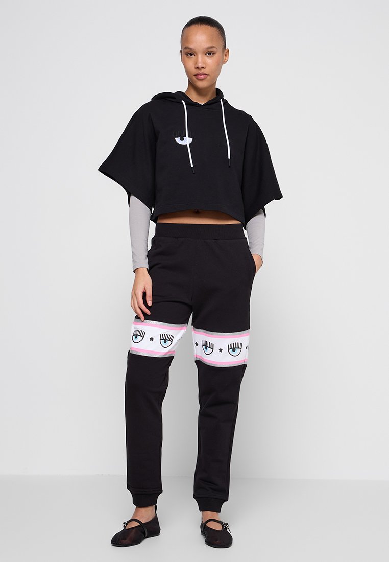 Schwarze, kurz geschnittene Kapuzen sweatshirt mit weißen Kordeln, kombiniert mit schwarzen Jogginghosen, die mit rosa und weißen grafischen Augendesigns verziert sind.