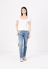 Blouse blanche à manches courtes avec un décolleté carré, associée à un jean ajusté bleu clair. Le modèle porte un sac à main blanc et des chaussures beiges.