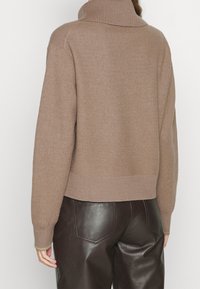 Brązowy, oversize'owy sweter z wysokim, prążkowanym kołnierzem. Miękka faktura, krótka długość i prążkowane mankiety. Połączony z ciemnymi, błyszczącymi skórzanymi spodniami.