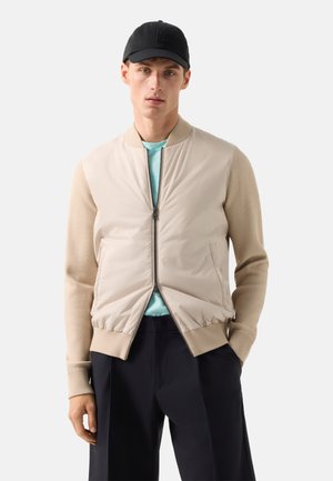 Mężczyzna w beżowej kurtce na zamek z dzianinowymi rękawami, jasnoniebieskiej koszuli, czarnych spodniach i czapce bejsbolowej, stojący na białym tle.