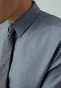 Graues Hemd mit Knopfleiste und Kragen, das eine glatte Textur aufweist und eine passende gewebte Krawatte hat, die teilweise auf der linken Seite sichtbar ist.