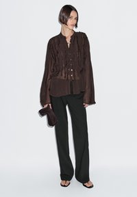 Brun bluse med flæser, knapper foran, transparent stof og lange ærmer, kombineret med sorte bukser med vide ben og en mørk clutch.