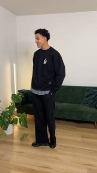 Sudadera negra oversize con logo, combinada con una camiseta gris claro, pantalones negros sueltos y zapatos negros voluminosos. Sofá verde de fondo.