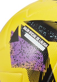 Geltonas futbolo kamuolys su tekstūruota paviršiumi, turintis juodus ir violetinius geometrinius raštus, ir etiketė, nurodanti „Oficialus LaLiga kamuolys 2024/2025 sezonui.“