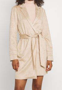 Cappotto beige in tessuto scamosciato, con colletto a scialle, vita con cintura e tasche laterali. Presenta dettagli di cucitura sulle maniche e lunghezza fino a metà coscia.
