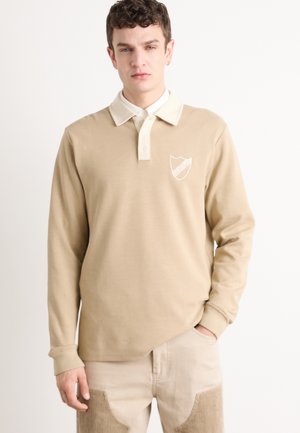 Beige Polohemd mit langen Ärmeln und einem kontrastierenden weißen Kragen, mit einem Wappen-Logo auf der linken Brust und einer glatten Stofftextur.