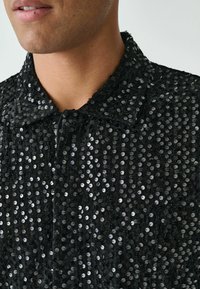 Schwarze Paillettenjacke mit einem Kragen. Der Stoff ist mit dicht aneinander gereihten, glänzenden runden Pailletten versehen, die eine strukturierte Optik erzeugen.