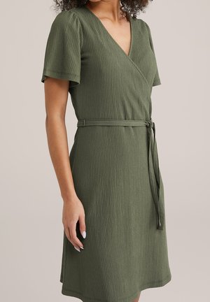 Kobieta ubrana w teksturowaną oliwkowozieloną sukienkę kopertową z krótkimi rękawami i zawiązanym paskiem w talii, pokazana od szyi do kolan na neutralnym tle.
