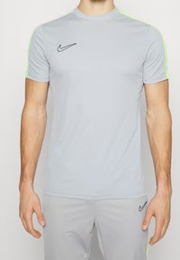Camiseta atlética gris de manga corta, con acentos en verde neón en los hombros y un logo negro de Nike en el pecho. Tejido elástico y ligero.