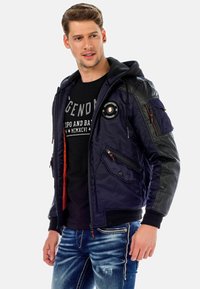 Dunkelblaues Bomberjackett mit schwarzen Lederakzenten, mit Reißverschluss versehenen Taschen und Kapuze. Verfügt über ein rundes Emblem auf der Brust und orangene Details.
