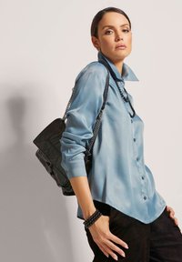 Lichtblauw satijnen shirt met knopen en lange mouwen, in combinatie met een zwarte tas met kettingriem en textuurelementen.