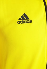 Keltainen polyesterikangas, jossa on sileä pinta, ja joka esittelee mustan Adidas-logon sekä mustan vetoketjun yksityiskohtana sivuilla.