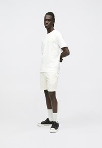 Hvit t-skjorte med korte ermer og grønt logo, kombinert med teksturerte lyse shorts. Sorte joggesko med hvite såler og hvite, mellomlange sokker.