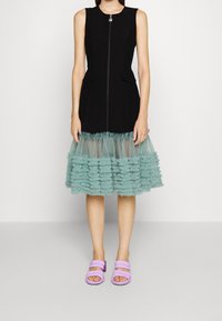 Robe noire sans manches avec une fermeture éclair, dotée d'un ourlet en tulle aqua à volants. Associée à des sandales plates lavande.