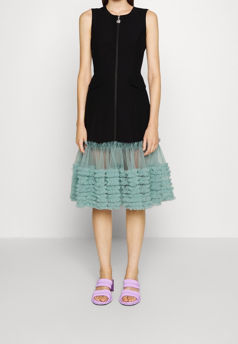 Robe noire sans manches avec une fermeture éclair, dotée d'un ourlet en tulle aqua à volants. Associée à des sandales plates lavande.