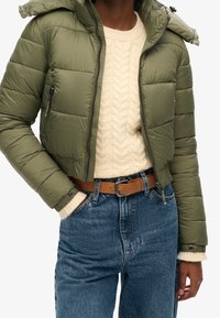 Oliwkowa puchowa kurtka z kapturem, pikowana, z zapięciem na zamek z przodu, noszona na kremowym swetrze w warkocze i ciemnoniebieskich dżinsach z wysokim stanem.