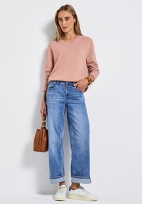 Pull v-neck rose clair, jeans bleu décontractés avec ourlets retroussés, baskets blanches et sac à main en cuir brun, mettant en avant une tenue décontractée.