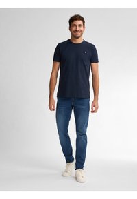Camiseta de algodón azul marino con cuello redondo y logo en el lado izquierdo del pecho, combinada con jeans azules y zapatillas blancas. Diseño sencillo y casual.