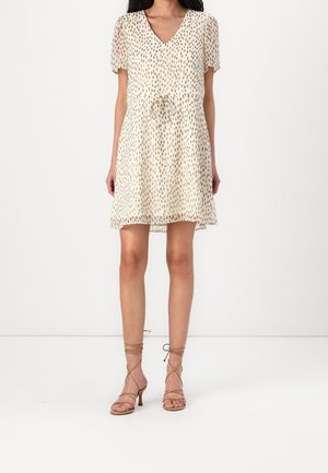 Mujer con vestido corto beige con patrón de puntos irregulares marrones, escote en V, mangas cortas y sandalias beige de tacón con cordones.