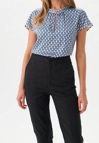 Femme portant un haut à manches courtes à motifs bleus et blancs rentré dans un pantalon tailleur noir taille haute.