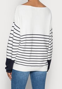 Kvinna iklädd en vit tröja med svarta horisontella ränder och svarta ärmslut, i kombination med blå denimjeans, sedd bakifrån.