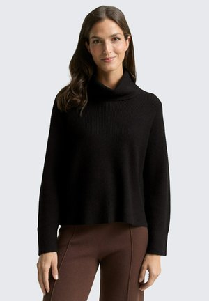 Black, ribbed knit sweater with a high cowl neck and long sleeves, paired with brown fitted pants. Soft texture, relaxed fit.