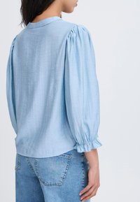 Ljusblå blus med puffärmar och elastiska manschetter, med en slät textur. Matchad med högmidjade denimjeans.