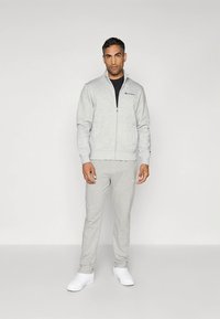 Pilkas džemperis su užtrauktuku, turintis būdingus rankogalius, derinamas su tokiomis pačiomis sportinėmis kelnėmis. Balti sportbačiai užbaigia aprangą. Tinka kasdieniam dėvėjimui.