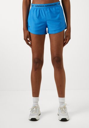 Vrouw draagt blauwe Under Armour sportshorts, witte crew-sokken en witte hardloopschoenen, staand tegen een effen achtergrond.