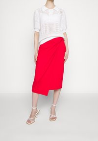 Rode wikkelrok met een knoopdetail, gecombineerd met een witte gebreide top met een kraag en korte poefmouwen. Wit gespt hakken.