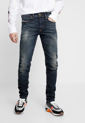 Homme portant un jean slim bleu foncé avec des détails délavés et des baskets noires, blanches et orange, debout devant un fond uni.