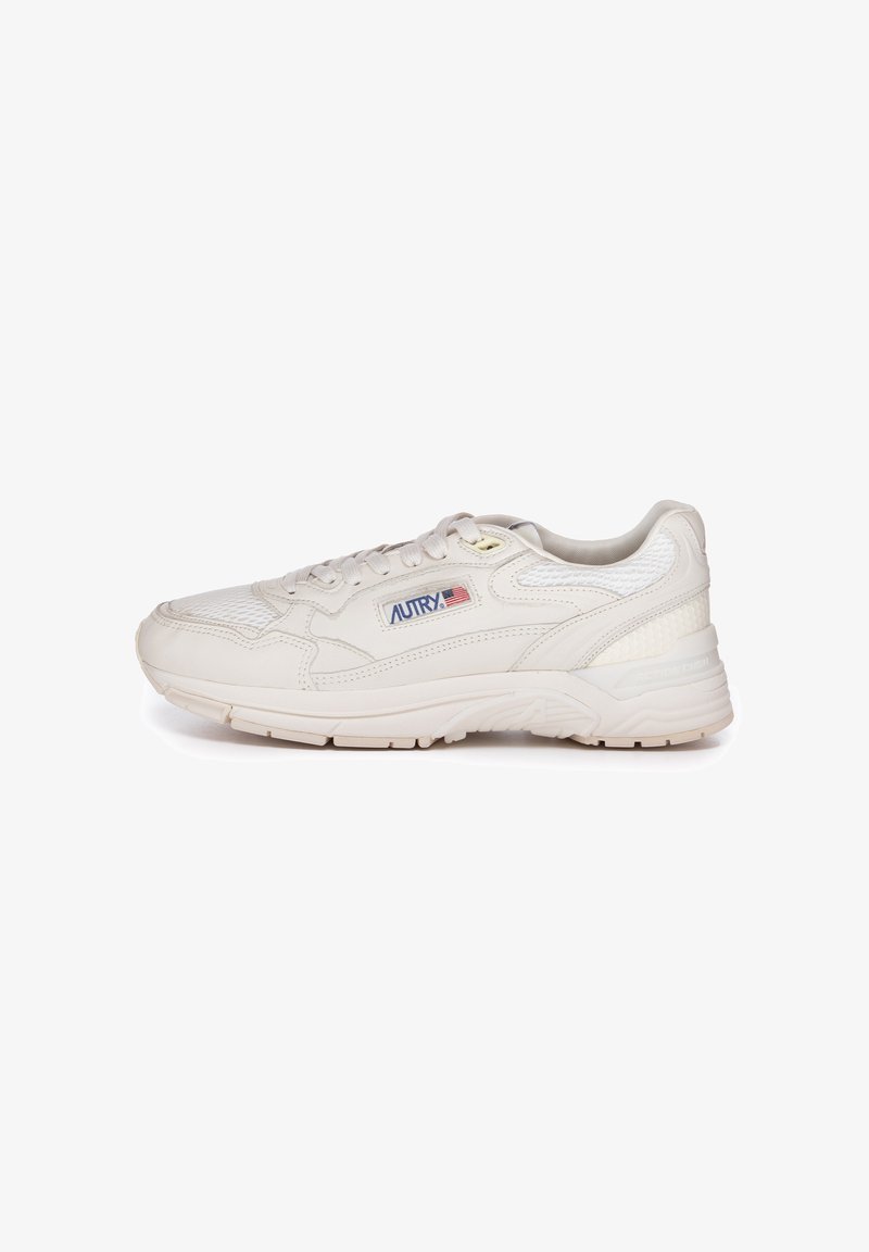 Zapatilla deportiva beige con parte superior de malla texturizada, cuello acolchado y suela de goma con patrones de relieve prominentes. Logo en el lateral.