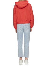 Rote, verkürzte Jacke mit Kapuze, elastischem Bund und langen Ärmeln, kombiniert mit hellblauen, geraden Jeans und weißen Sneakers.
