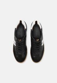 Fekete velúr és bőr sneaker cipők fehér csíkkal és arany logóval. Fűzős kialakítás, párnázott talpbetét és gumiból készült talp.