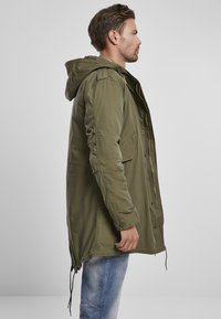 Olivově zelená parka s kapucí, vybavená zipem vpředu, šňůrkami na spodním lemu a bočními kapsami. Vyrobena z lehkého materiálu.