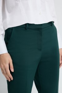 Pantaloni verdi a sigaretta con una texture liscia, caratterizzati da cuciture frontali e tasche laterali, abbinati a una camicia bianca corta con colletto.