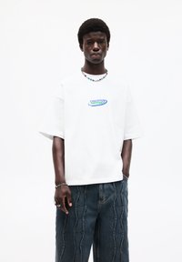 White oversized T-shirt with "CREATIVE MINDSET" graphic in blue and green. Paired with dark blue wide-leg jeans featuring wavy stitch detailing.