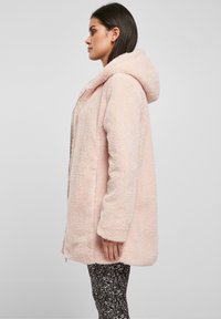 Giacca cappuccio in pelliccia artificiale rosa con vestibilità rilassata e zip frontale, caratterizzata da una texture morbida e maniche lunghe. Abbinata a leggings neri con fantasia.